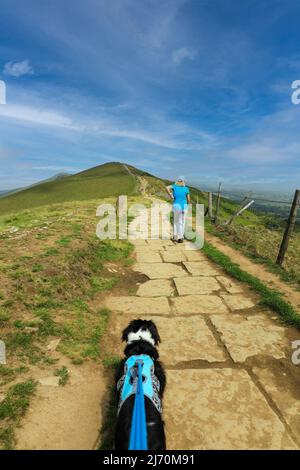 Eine Wanderin und ein Hund auf einem asphaltierten Pfad auf dem Gipfel des Mam Tor Castleton im High Peak of Derbyshire, Peak District National Park, England, Großbritannien Stockfoto