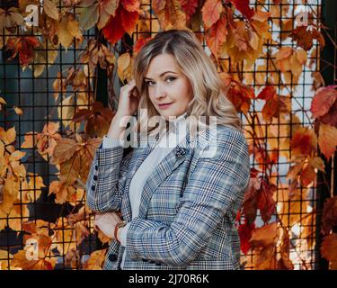 Schöne Frau in der Nähe von Kletterpflanzen auf Zaun mit roten und gelben Blättern im Herbst Stadtpark Nahaufnahme. Erwachsene Dame, die auf der Straße spazierenging und sich erholte Stockfoto