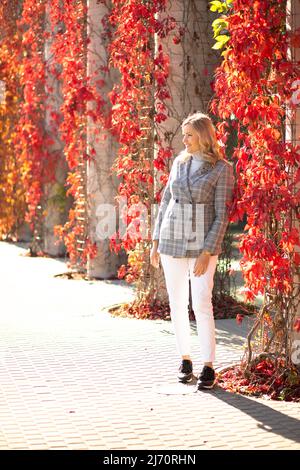 Glückliche Frau in der Nähe von Kletterpflanzen mit roten und gelben Blättern im Herbst Stadtpark an sonnigen Tagen. Lächelnde Dame, die auf der Straße spazierenging und sich in der Stadt erholte Stockfoto