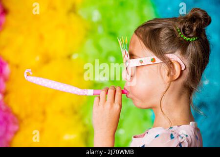 Süßes Mädchen feiert Geburtstag, trägt Partybrille, bläst Partyhörner, lacht und hat Spaß. Heller regenbogenfarbener Hintergrund. Glücklich Stockfoto