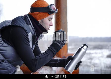 Ein Mann in einem orangenen Hut im Winter trinkt Tee aus einer Thermoskanne während eines Winterurlaubs in den Bergen. Er steht auf dem Balkon eines Holzhauses. Anschluss Stockfoto