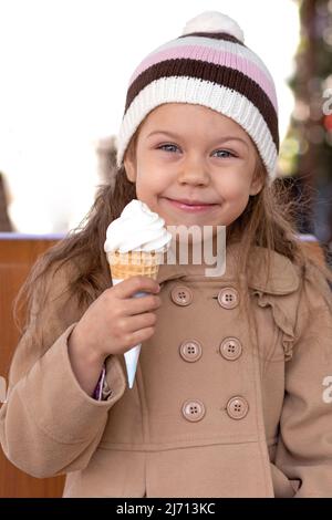 Porträt eines glücklichen und lächelnden kaukasischen kleinen Mädchens von 5-6 Jahren, das draußen Eiscreme-Kegel in hellbraunem Mantel und Hut hält Stockfoto
