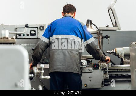 Turner steht hinter der Drehmaschine in der Produktionshalle und arbeitet. Blick auf den Arbeiter von hinten in Overalls. Authentische Arbeitsprozess-Szene in der Produktion. Echter Arbeiter. Nicht inszenierte Szene. Stockfoto