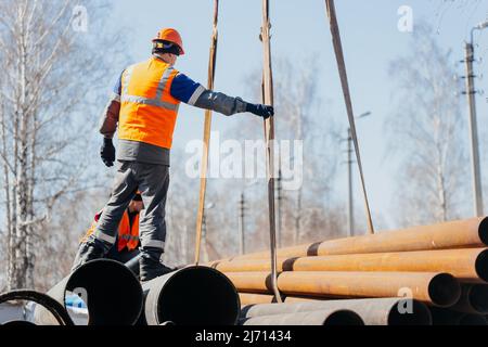 Slinger im Helm entlädt am sonnigen Sommertag Metallrohre auf der Baustelle. Echte Szene. Workflow. Echter Arbeiter. Stockfoto