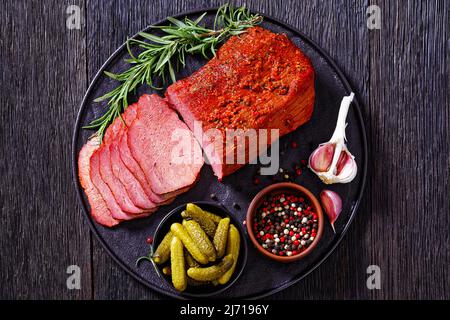 Frisch geschnittenes Rinderpastrami auf schwarzem Teller mit frischem Rosmarin, Knoblauch und Gurken auf dunklem Tisch, horizontale Ansicht von oben, flach liegend Stockfoto
