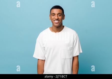 Porträt eines fröhlichen jungen afroamerikanischen Mannes, der ein weißes, legeres T-Shirt trägt, das isoliert auf blauem Hintergrund posiert. Sorgloser junger Mann, der die Kamera anschaut und lacht Stockfoto