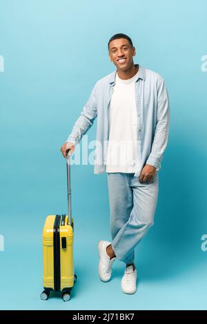 In voller Länge Foto von lächelnden jungen afroamerikanischen Kerl in legerer Denim-Kleidung stehen mit gelben Koffer isoliert auf blauem Hintergrund, Reise-und Urlaubskonzept Stockfoto