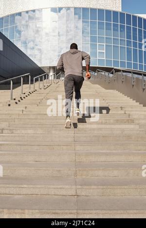 Der nicht zu erkennende, sportliche Schwarze, der eine Betontreppe zum modernen Business Center hinaufläuft, während er im Freien trainieren und dabei den Blick auf die Rückseite aus der Nähe schweissen lässt Stockfoto