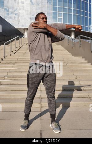 Der junge afroamerikanische Sportler im aktiven Sport macht Aufwärmübungen, während er im Freien vor dem modernen Business Center steht Stockfoto
