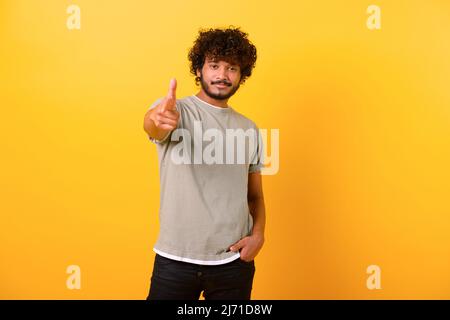 Porträt eines jungen indischen Mannes, der ein legeres T-Shirt trägt, mit der Fingerpistole spielerisch auf die Kamera zeigt und flirtet und den glücklichen Gewinner wählt. Innenaufnahme des Studios isoliert auf gelbem Hintergrund Stockfoto
