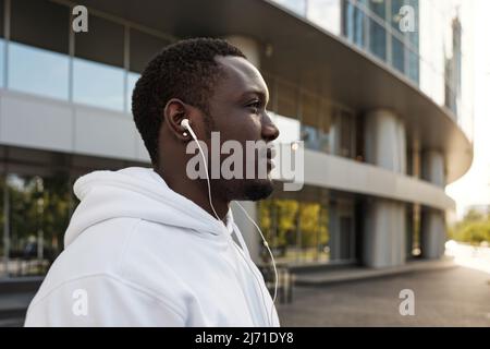 Profilportrait eines jungen afroamerikanischen Mannes in weißem Hoodie und Ohrhörer, der im Freien in die Ferne blickt Stockfoto