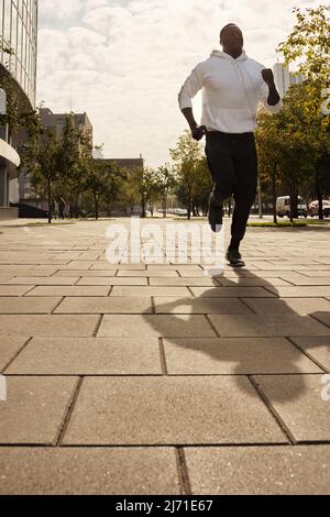 Blick aus der Perspektive auf einen jungen, athletischen Afroamerikaner in Sportkleidung, der am sonnigen Morgen auf dem Bürgersteig in der Innenstadt läuft Stockfoto