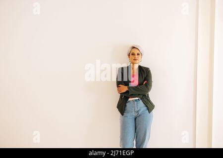 Geschäftsfrau, die in die Kamera blickt, während sie mit überkreuzten Armen steht. Selbstbewusste junge Geschäftsfrau, die vor einem Wandhintergrund in einem modernen Stockfoto