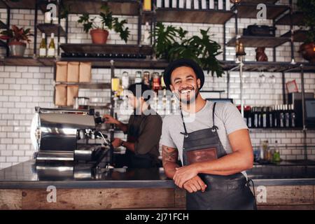 Mitarbeiter im Café lächelt vor der Kamera und steht an der Theke. Fröhlicher junger Mann auf der Schürze und dem Hut, der sich zum Cafe-Tresen lehnte, während der Kellner im Hintergrund arbeitete Stockfoto