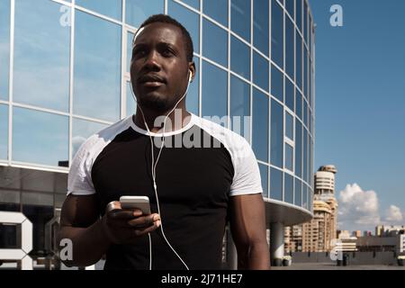 Blick aus der unteren Perspektive auf einen hübschen afrikanischen Mann, der mit einem Smartphone gegen ein modernes Geschäftsgebäude steht und über Kopfhörer Musik hört Stockfoto