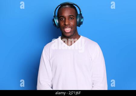 Porträt des Millennials Mann trägt Kopfhörer, lächelt breit, genießt Lieblingsmusik, gekleidet in weißen Pullover, isoliert auf blauem Hintergrund, hat glücklich Stockfoto