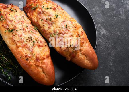 Baguette-Boote. Heiß gebackenes Sandwich auf Baguette-Brot mit Schinken, Speck, Gemüse und Käse auf Pergament und schwarzem dunklen Betonboden. Draufsicht. Stockfoto