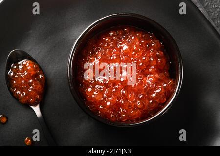 Kaviar mit rotem Lachs. Köstlicher roter Kaviar in schwarzer Schale auf schwarzem dunklen Betongrund. Draufsicht mit Kopierbereich. Flach liegend. Modell. Stockfoto