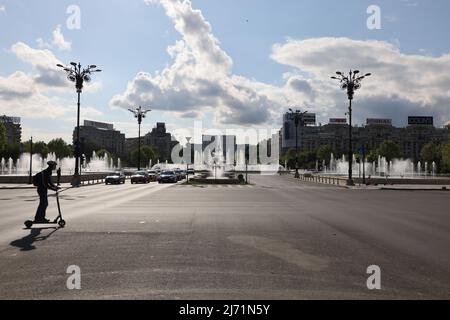 Leerer Platz mit Springbrunnen und Blick auf das Parlamentsgebäude, während eine Person mit Helm an einem Elektroroller vorbeifährt; sonnig mit Wolken am Himmel Stockfoto