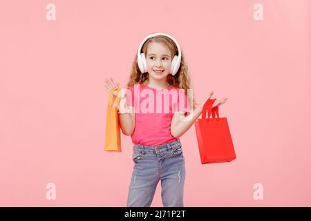 Liebenswert Kind Mädchen in Kopfhörer hält Einkaufstaschen in den Händen über dem Kopf isoliert auf rosa Hintergrund. Shopping-Konzept. Stockfoto