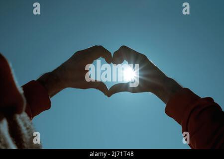 Herzform machen von Händen gegen hellen Tag. Zwei Hände bilden eine herzförmige Silhouette am Himmelskreuz. Stockfoto