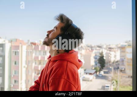 Friedlicher Mann in modernen kabellosen Kopfhörern, der auf dem Balkon steht und Musik hört, glücklicher, ruhiger junger Mann in Ohrhörern, der sich ausruht. Genießen Sie guten Sound, Stockfoto