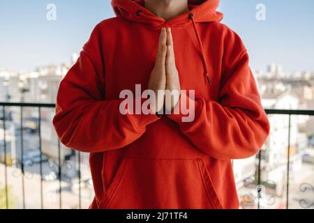 Friedlicher, religiöser junger Mann mit rotem Hoodie, der die Hände über der Brust mit geschlossenen Augen hält. Er faltete die Handflächen bei h Stockfoto