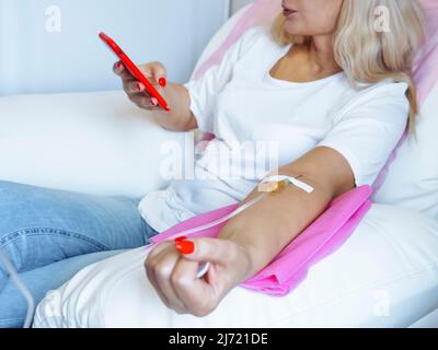 Spenderin schaut auf das Mobiltelefon, während die Nadel im Arm ist, Nahaufnahme des Arms mit der Nadel in den Arm eingeführt Stockfoto