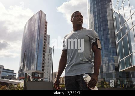 Blick aus der Nähe auf den athletischen afroamerikanischen Mann mit Ohrhörern und Mobiltelefon im Armband, der in der Innenstadt steht Stockfoto