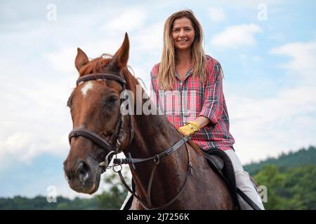 Junge Frau im Hemd, die sich auf einem braunen Pferd ausruht - sein Fell ist vom Schweiß nass - nach dem Reiten, lächelnd Stockfoto