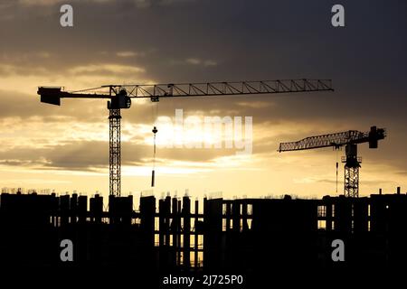 Silhouetten von Turmdrehkranen und Arbeitern auf Gerüsten von unfertigen Gebäuden bei Sonnenaufgang. Wohnungsbau, Wohnblock in der Stadt auf dem Hintergrund Stockfoto