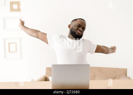 Der afroamerikanische Mann, der am Laptop arbeitet, leidet unter Rückenschmerzen und verstauchert seinen Rücken, nachdem er lange Zeit zu Hause am Laptop gearbeitet hat Stockfoto