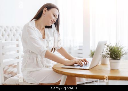 Frau, die in ihrem Schlafzimmer mit dem Mobiltelefon spricht und einen Laptop benutzt. Stockfoto