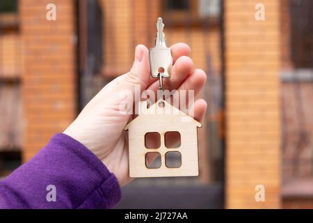 Hand hält einen Schlüssel mit einem flachen hölzernen Schlüsselanhänger im Freien. Wohnimmobilieninvestitionskonzept. Stockfoto