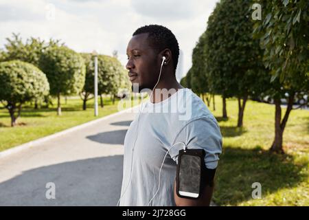 Porträt eines schönen, muskulösen afroamerikanischen Läufers mit Ohrhörern und Smartphone im Armband, der im Park steht und sich ausruht Stockfoto