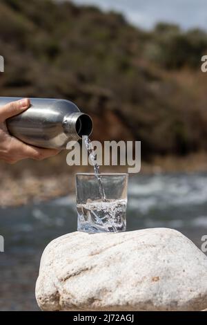 Nahaufnahme Gießen gereinigtes frisches Trinkwasser aus der Flasche auf Glas Tasse in der Natur mit Fluss hinter Stockfoto