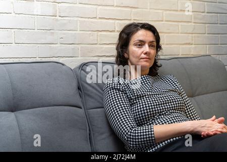 Eine europäische Frau mittleren Alters mit dunklen Haaren sitzt auf einem Sofa Stockfoto