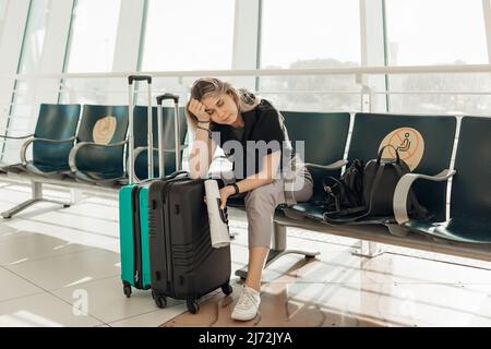Gelangweilte blonde Frau mit Gepäck, Ellenbogen auf Taschen lehnend, sitzen im Wartezimmer am Flughafen wegen Reisebeschränkungen von Covid Stockfoto
