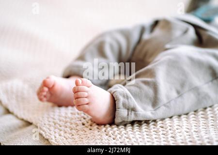 Unbekanntes barfuss-Kleinkind mit grauem Body auf beigem Baumwollgestrick. Nahaufnahme von wunderschönen winzigen Babyfüßen auf dem Bett. Schlafenszeit Stockfoto