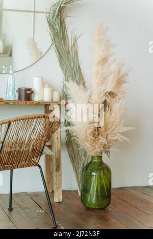 Einrichtung eines modernen, hellen und stilvollen Zimmers. Großer runder Spiegel an weißer Wand. Enger Holztisch mit verschiedenen Kerzen, Glasflasche in der Nähe von Korbstuhl. Hu Stockfoto