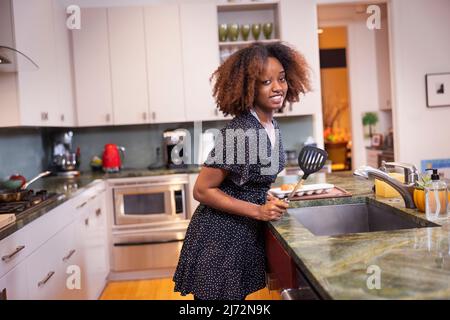 Junge afroamerikanische Frau, die einen Spachtel in der Hand hält, während sie in einer Wohnküche kocht Stockfoto