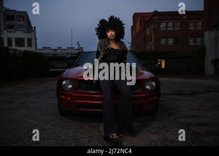 Afroamerikanische Frau, die am Abend stolz auf der Motorhaube ihres Autos posiert. Stockfoto