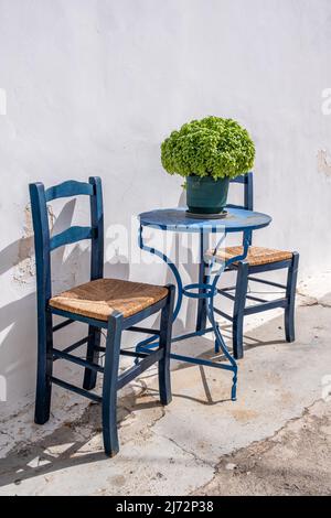 Leerer blauer runder Metalltisch und zwei Holzstühle, weiße traditionelle Gebäudewand. Café im Freien auf einer griechischen Insel Chora, Griechenland. Sommersonntag. Stockfoto