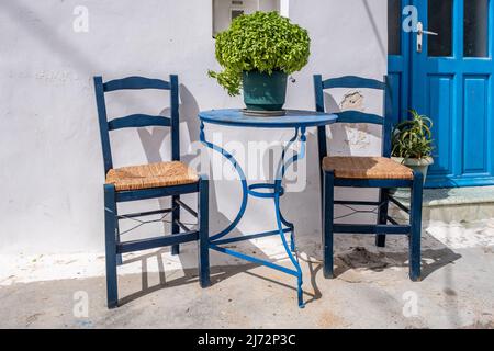 Leerer blauer runder Metalltisch und zwei Holzstühle, weiße traditionelle Gebäudewand. Café im Freien auf einer griechischen Insel Chora, Griechenland. Sommersonntag. Stockfoto