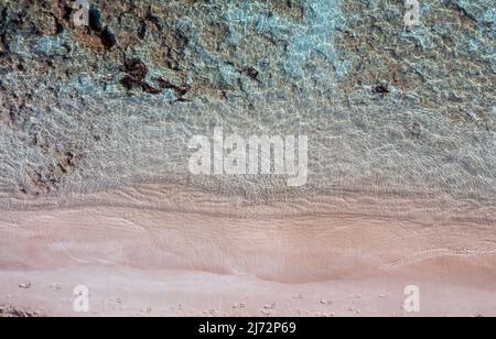 Sandstrand, türkisblaues Meer Luftaufnahme von oben. Fußabdrücke auf nassem rosa Sand. Klares, transparentes Wasser, felsiger Meeresboden. Ägäische griechische Insel Sum Stockfoto