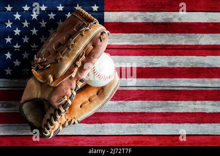 Leder-Baseballhandschuh oder Softballhandschuh mit Ball auf rustikalem Holzhintergrund mit bemalter US-Flagge und Kopierraum. Stockfoto
