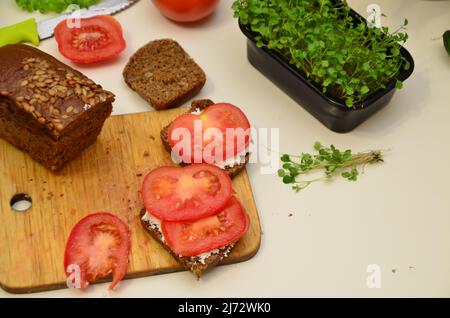 Das Mädchen kocht und isst zum Frühstück Salat, Tomate, Weichkäse. Veganes Sandwich mit Roggenbrot, Spinat, mikrogrün isoliert auf weiß, zu Stockfoto