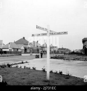 Cliftonville, Kent. 1950er Jahre - Ein neues Wegweiser, das für das öffentliche Haus „YE Olde Charles Inn“, Cliftonville, Margate, Kent errichtet wurde. Das Schild zeigt auch Wegbeschreibungen und Entfernungen nach Moskau, Monte Carlo und Grönland. Ein Straßenkehrer schaut in der Ferne an. Stockfoto