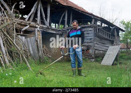 Mann mäht hohes Gras mit Benzin Rasentrimmer im Dorf rustikalen Hof. Gartengeräte. Prozess des Rasentrimmens mit dem Handmäher Stockfoto