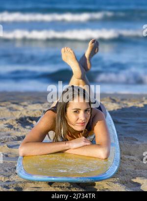 Junge Frau, die auf einem Surfbrett auf dem Sand liegt und geradeaus blickt Stockfoto